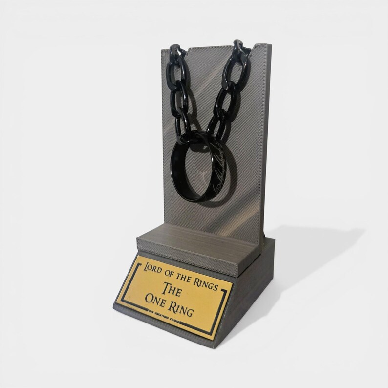 May include: A black ring and chain necklace displayed on a gray stand. The stand has a gold plaque with the text "Lord of the Rings The One Ring". The ring has an inscription. The chain is made of large, oval links.