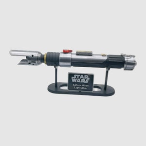 Pode incluir: Um sabre de luz prateado e preto com um bot&atilde;o vermelho na empunhadura, exibido em um suporte preto com uma placa prateada que diz "STAR WARS Sabine Wren Lightsaber".
