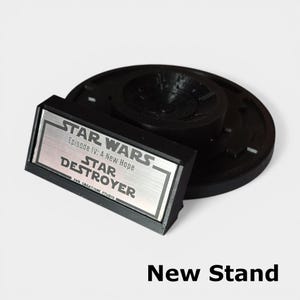 May include: A black Star Destroyer stand with a silver nameplate. The nameplate reads "STAR WARS Episode IV: A New Hope STAR DESTROYER". The stand is round with a central support and the text "New Stand" is at the bottom.