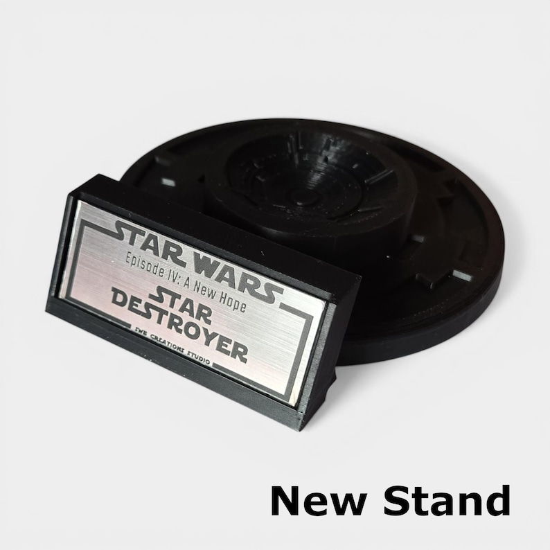 May include: A black Star Destroyer stand with a silver nameplate. The nameplate reads "STAR WARS Episode IV: A New Hope STAR DESTROYER". The stand is round with a central support and the text "New Stand" is at the bottom.
