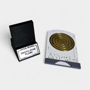 May include: A black stand with a silver plaque that reads "STAR WARS DEATH STAR PLANS". Next to the stand is a white plastic replica of a data card with a gold disc inside.