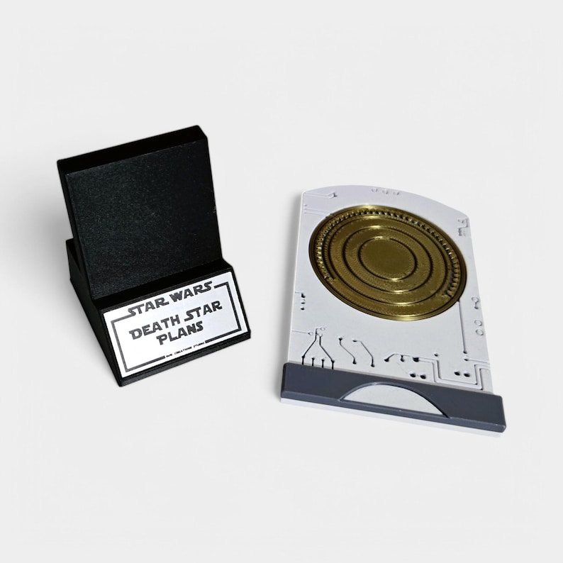 May include: A black stand with a silver plaque that reads "STAR WARS DEATH STAR PLANS". Next to the stand is a white plastic replica of a data card with a gold disc inside.
