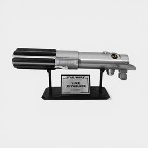 May include: A silver and black lightsaber replica, displayed on a black stand. The lightsaber has a detailed handle and a prominent emitter. A plaque on the stand reads "Luke Skywalker - Lightsaber" with Star Wars branding.