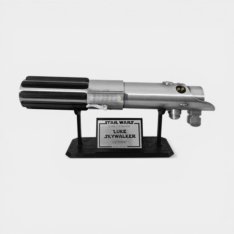 May include: A silver and black lightsaber replica, displayed on a black stand. The lightsaber has a detailed handle and a prominent emitter. A plaque on the stand reads "Luke Skywalker - Lightsaber" with Star Wars branding.