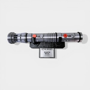Może przedstawiać: Srebrna replika miecza świetlnego Darth Maula, zamontowana na czarnym stojaku. Miecza świetlny ma czerwone przyciski i szczeg&oacute;łowy uchwyt. Poniżej tabliczka Star Wars z napisem "Darth Maul Lightsaber."