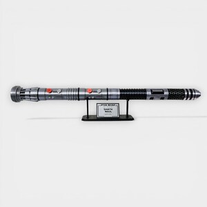 Puede incluir: R&eacute;plica de sable de luz plateado y negro con botones rojos, exhibida en un soporte negro. El soporte tiene una etiqueta que dice "Star Wars Darth Maul Lightsaber". El sable de luz es un accesorio detallado.