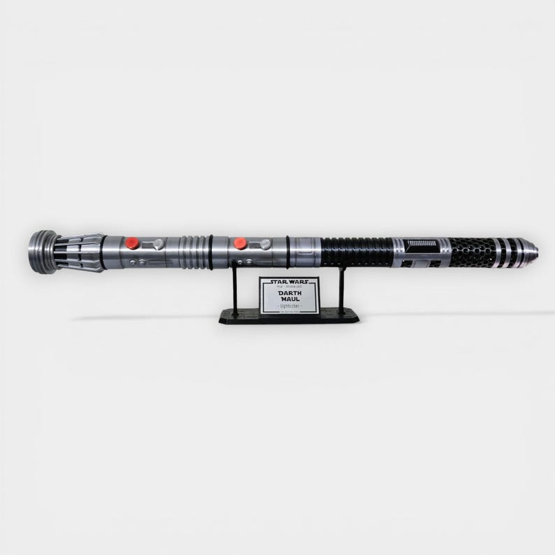 Puede incluir: R&eacute;plica de sable de luz plateado y negro con botones rojos, exhibida en un soporte negro. El soporte tiene una etiqueta que dice "Star Wars Darth Maul Lightsaber". El sable de luz es un accesorio detallado.