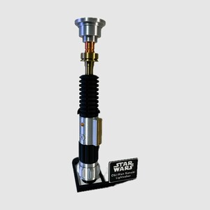 May include: A silver, gold, and black lightsaber replica standing vertically on a black display stand. The hilt features a ribbed black grip, a metallic silver emitter, and a small rectangular plaque reading STAR WARS Obi-Wan Kenobi Lightsaber.