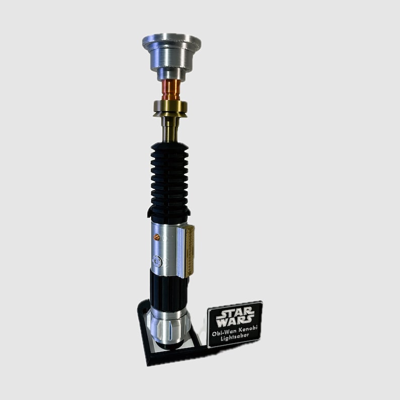 May include: A silver, gold, and black lightsaber replica standing vertically on a black display stand. The hilt features a ribbed black grip, a metallic silver emitter, and a small rectangular plaque reading STAR WARS Obi-Wan Kenobi Lightsaber.