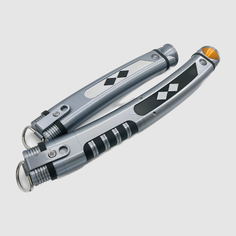 Peut inclure: Deux accessoires cylindriques argent&eacute;s aux corps incurv&eacute;s, dot&eacute;s de sections de pr&eacute;hension noires, d'incrustations m&eacute;talliques en forme de losange et d'un petit accent triangulaire orange &agrave; l'extr&eacute;mit&eacute;. Chaque pi&egrave;ce inclut un anneau m&eacute;tallique.