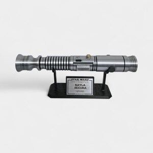 May include: A silver lightsaber prop, displayed on a black stand. The prop has a detailed design with various textures and a button. A plaque on the stand reads "Aayla Secura Lightsaber" with the Star Wars logo.