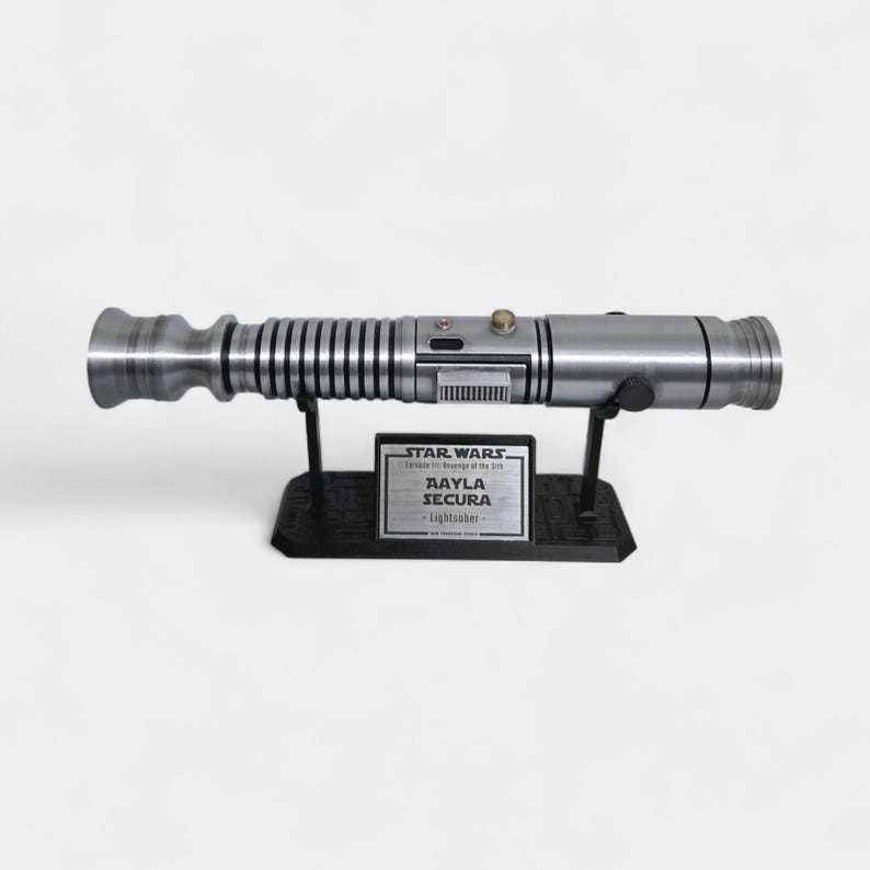 May include: A silver lightsaber prop, displayed on a black stand. The prop has a detailed design with various textures and a button. A plaque on the stand reads "Aayla Secura Lightsaber" with the Star Wars logo.