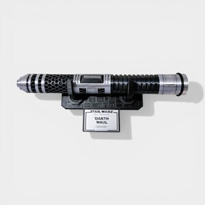 Puede incluir: R&eacute;plica del sable de luz de Darth Maul, con dise&ntilde;o plateado y negro. El sable de luz est&aacute; montado en un soporte negro con una placa que dice "Star Wars - Darth Maul - Lightsaber".