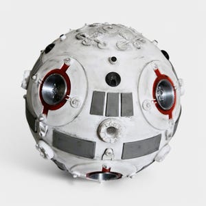 May include: A white and gray spherical robot with red accents and multiple circular details. The robot has a textured surface and appears to be made of metal.