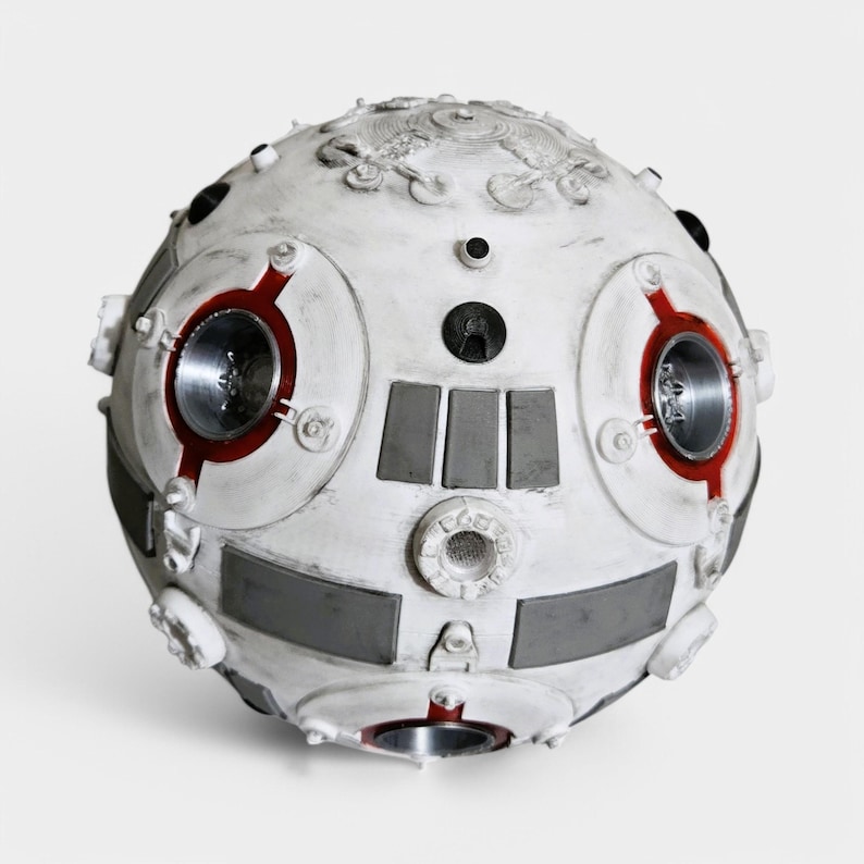 May include: A white and gray spherical robot with red accents and multiple circular details. The robot has a textured surface and appears to be made of metal.