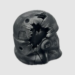 K&ouml;nnte beinhalten: Ein schwarzer, zerbrochener Plastikhelm mit einer rissigen und besch&auml;digten Oberfl&auml;che. Der Helm hat ein Visier und ein kleines, rundes, schwarzes Objekt an der Seite.