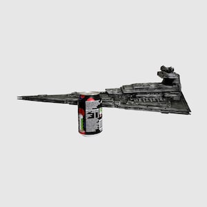 May include: A detailed, dark gray model of a Star Destroyer spaceship, with intricate surface details. A can of soda with a black, red, and white design is placed next to the model for scale. The spaceship model is a collectible item.