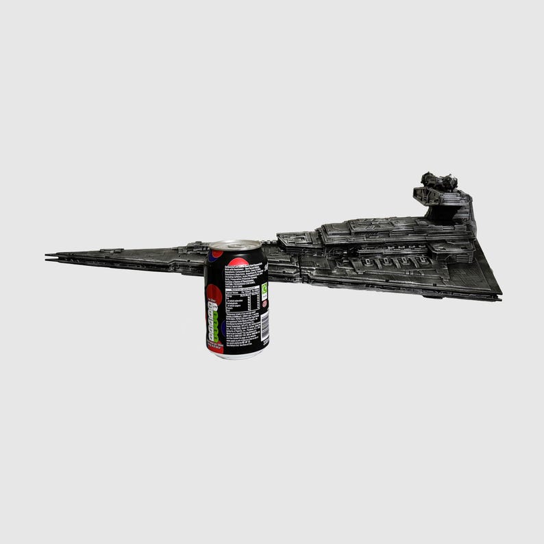 May include: A detailed, dark gray model of a Star Destroyer spaceship, with intricate surface details. A can of soda with a black, red, and white design is placed next to the model for scale. The spaceship model is a collectible item.