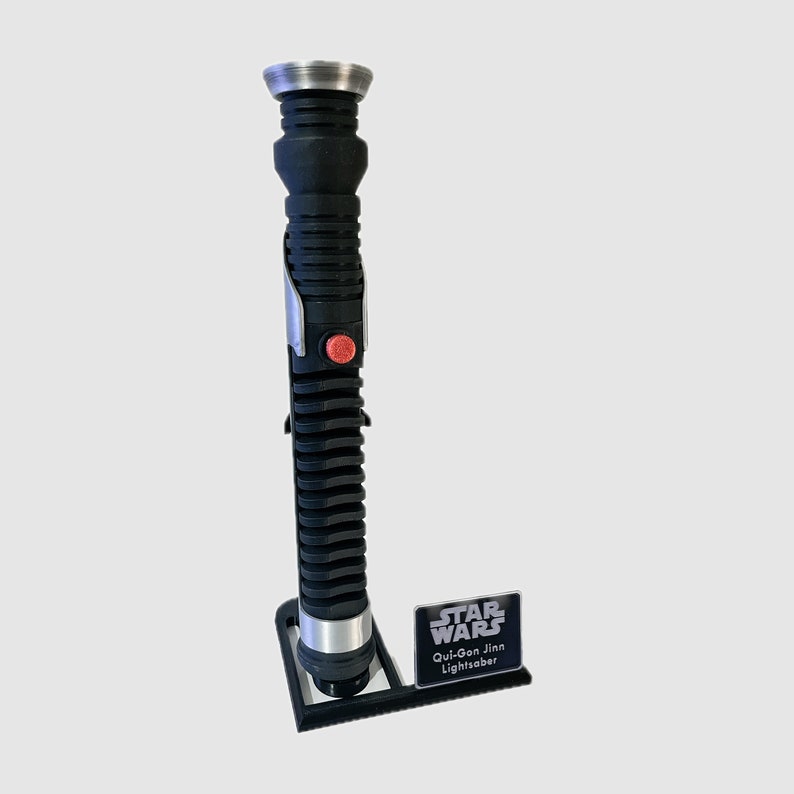 May include: A black lightsaber with a silver handle and a red button. The lightsaber is on a black stand with a silver "Star Wars" logo and the text "Qui-Gon Jinn Lightsaber".