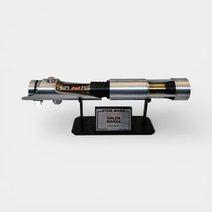 May include: A silver and black lightsaber replica, displayed on a black stand. The lightsaber features intricate internal details and is labeled "GALEN MAREK Lightsaber" with the Star Wars logo.