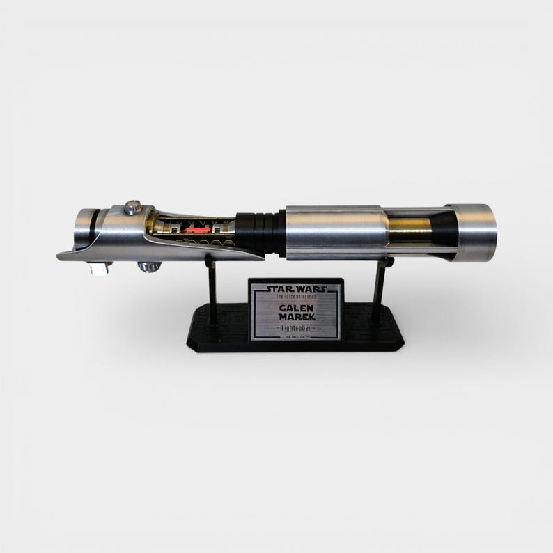 May include: A silver and black lightsaber replica, displayed on a black stand. The lightsaber features intricate internal details and is labeled "GALEN MAREK Lightsaber" with the Star Wars logo.