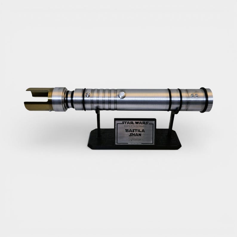 May include: A silver lightsaber prop from Star Wars, resting on a black stand. The lightsaber has a metallic finish with gold accents on the blade emitter. The stand features a plaque that reads "BASTILA SHAN Lightsaber".