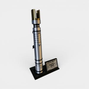 May include: A silver and gold lightsaber prop with a black base and a nameplate that reads "STAR WARS, BASTILA SHAN, lightsaber". The lightsaber has a metallic finish with detailed ridges and a gold emitter.