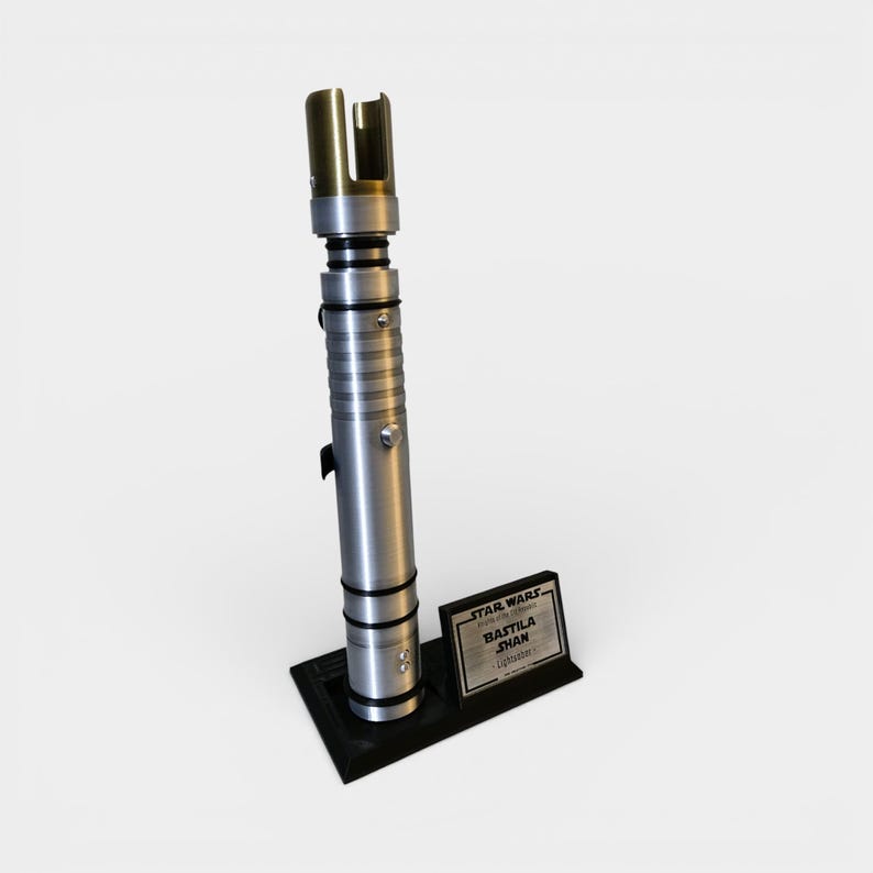 May include: A silver and gold lightsaber prop with a black base and a nameplate that reads "STAR WARS, BASTILA SHAN, lightsaber". The lightsaber has a metallic finish with detailed ridges and a gold emitter.