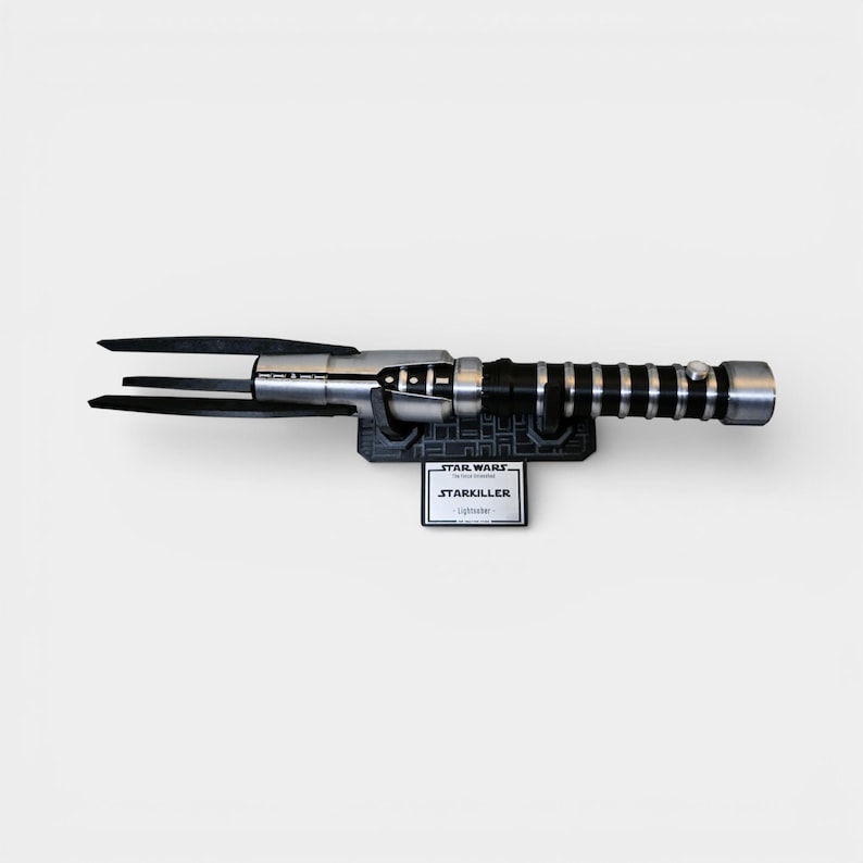 Op de afbeelding: Een Star Wars Starkiller lichtzwaard replica. Het lichtzwaard heeft een zwart en zilveren handvat met een geribbelde grip en een drietandig blad. Het is gemonteerd op een zwarte basis met een Star Wars logo en een Starkiller naamplaatje.