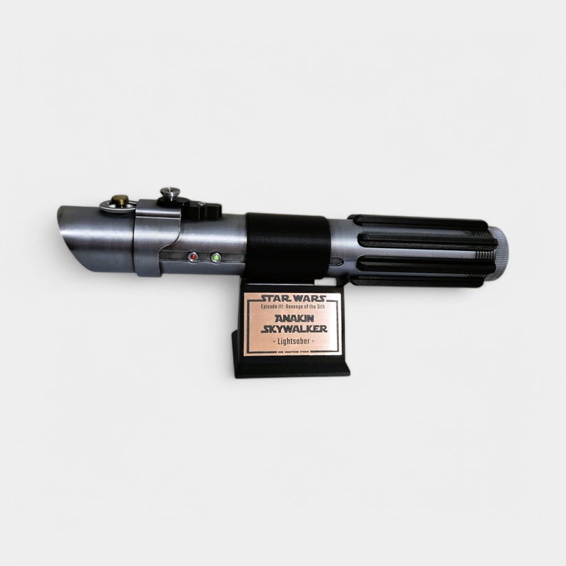 May include: A silver and black lightsaber replica from Star Wars: Episode III Revenge of the Sith. The lightsaber is on a stand with a plaque that reads "Star Wars Episode III Revenge of the Sith Anakin Skywalker - Lightsaber - "