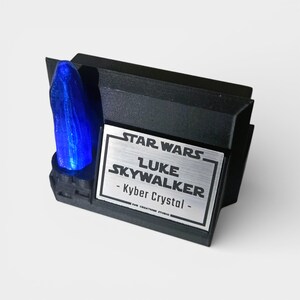 May include: A black display stand featuring a blue Kyber Crystal. The stand has a silver plaque with the Star Wars logo and the text "LUKE SKYWALKER - Kyber Crystal". The display is a collectible item.