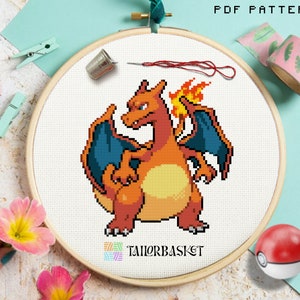 Może przedstawiać: Wzór do haftu krzyżykowego przedstawiający Charizarda, Pokemona w kolorze czerwonym, pomarańczowym i żółtym, z niebieskimi skrzydłami i płomiennym ogonem. Wzór znajduje się na białym tle i jest otoczony drewnianym obręczą do haftu.