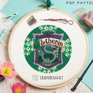Puede incluir: Un patrón de punto de cruz para un escudo de la casa Slytherin de la serie Harry Potter. El escudo es verde, morado y plateado con una serpiente y las palabras "Slytherin" en una pancarta.