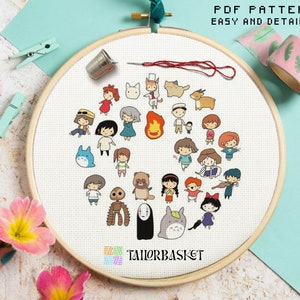 May include: A white embroidery hoop with a cross-stitch pattern of characters from Studio Ghibli films. The characters are arranged in a circle around a heart shape. The hoop is on a light blue surface.