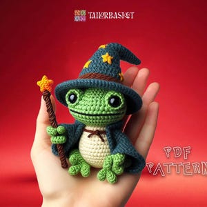 Puede incluir: Una rana de crochet verde que lleva un sombrero de mago azul con estrellas amarillas. La rana sostiene una varita marrón y naranja con una estrella amarilla en la parte superior. La rana está sentada en una mano, y el fondo es rojo. PATRÓN PDF.