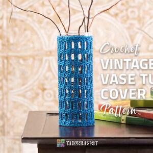 May include: A blue crocheted vase cover with a square pattern. The cover is on a glass vase with dried branches inside. The vase is on a wooden table with a stack of books.