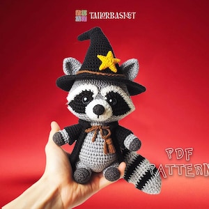May include: A crocheted raccoon wearing a witch hat with a yellow star. The raccoon is holding a brown and gold tassel. The raccoon is sitting on a hand.