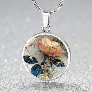 May include: A silver pendant necklace featuring a round charm with a floral design. The charm showcases a pink and gold rose with blue leaves against a white background with gold accents. The necklace has a silver chain.