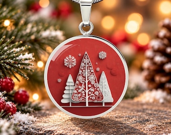 Op de afbeelding: Een ronde zilveren hanger met een rode achtergrond en drie gestileerde witte kerstbomen. De middelste boom heeft ingewikkelde sneeuwvlokpatronen, terwijl de omliggende bomen effen wit zijn. Kleine witte sneeuwvlokken zweven erboven.