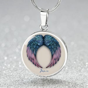 Puede incluir: Collar de plata con un colgante redondo que presenta un diseño de alas de ángel azul y rosa. El colgante tiene el nombre "Jamie" inscrito.