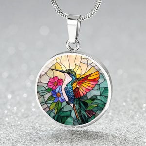 Puede incluir: Collar con colgante plateado con un colgante redondo con un diseño de colibrí colorido. El colibrí tiene alas azules, verdes, amarillas y rojas, con flores rosas y azules. El colgante cuelga de una cadena plateada.