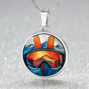 May include: A silver pendant necklace with a round charm featuring a ski goggles design. The goggles are orange and blue, with a mountain and sunset scene inside. Two orange skis are above the goggles. The necklace has a silver chain.