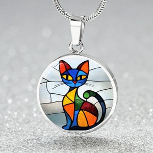 Può includere: Una collana con catena d'argento con un ciondolo rotondo che presenta un design di gatto astratto colorato. Il gatto è blu, arancione, giallo, verde e rosso.