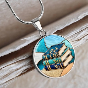 May include: A silver-toned circular pendant necklace featuring a graphic illustration of three stacked books with gold leaf accents against a blue and teal stained glass style background, suspended from a matching silver snake chain.