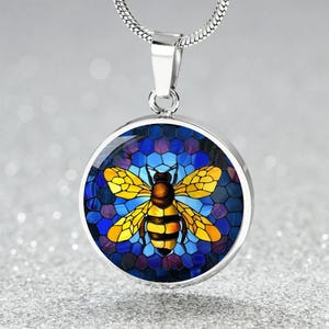 May include: A silver-toned pendant necklace featuring a round charm with a bee design. The bee has yellow wings and a brown body, set against a blue and purple honeycomb background. The necklace has a silver chain.