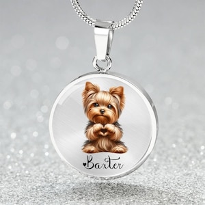 Op de afbeelding: Zilveren ketting met een ronde hanger met een cartoon Yorkshire Terrier met bruin en zwart haar. De hond houdt zijn poten bij elkaar voor zijn borst. De hanger heeft de naam "Baxter" onder de hond.