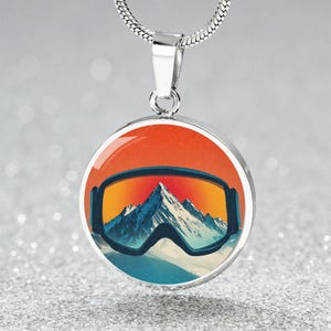 May include: Silver-toned pendant necklace featuring a round pendant with a ski goggles design. The pendant showcases a mountain scene with orange, yellow, and blue hues. The necklace has a silver chain.