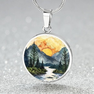 May include: A silver necklace with a round pendant featuring a watercolor painting of a mountain landscape with a yellow sun setting behind the mountains. The pendant is surrounded by a silver border.