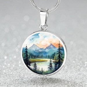 May include: A silver-toned circular pendant necklace featuring a colorful landscape illustration of snow-capped mountains, a calm lake, and dark evergreen trees under a soft sky with small birds flying in the distance, suspended on a silver chain.