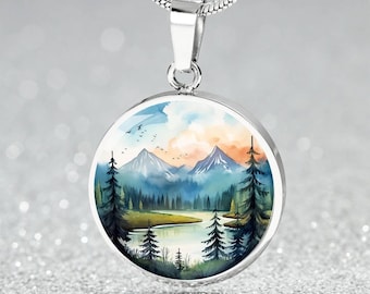 May include: A silver-toned circular pendant necklace featuring a colorful landscape illustration of snow-capped mountains, a calm lake, and dark evergreen trees under a soft sky with small birds flying in the distance, suspended on a silver chain.